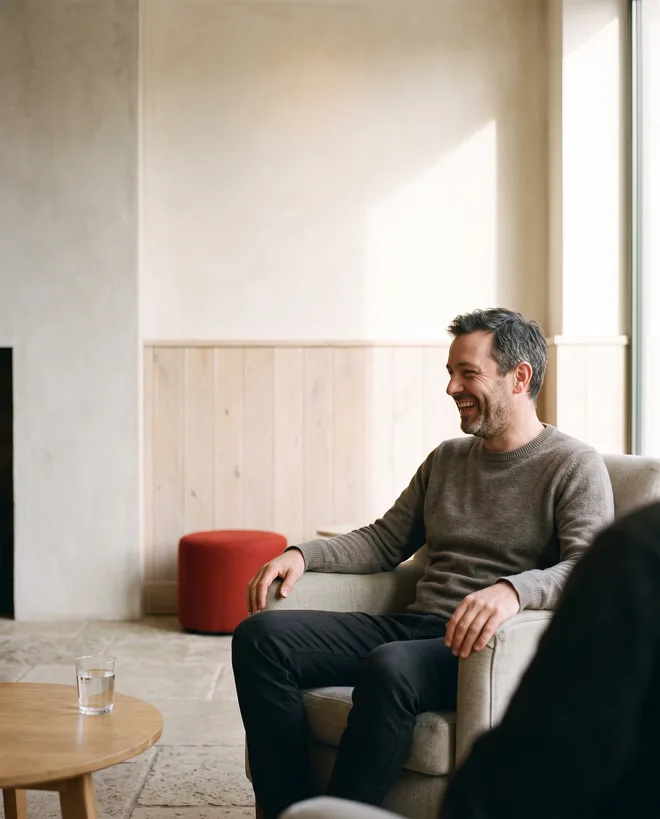 A copywriter laughing in a low armchair in a sunlit hotel lobby, hands resting on the chair arms, a companion only a soft shoulder at the edge of the frame and a red upholstered stool behind him