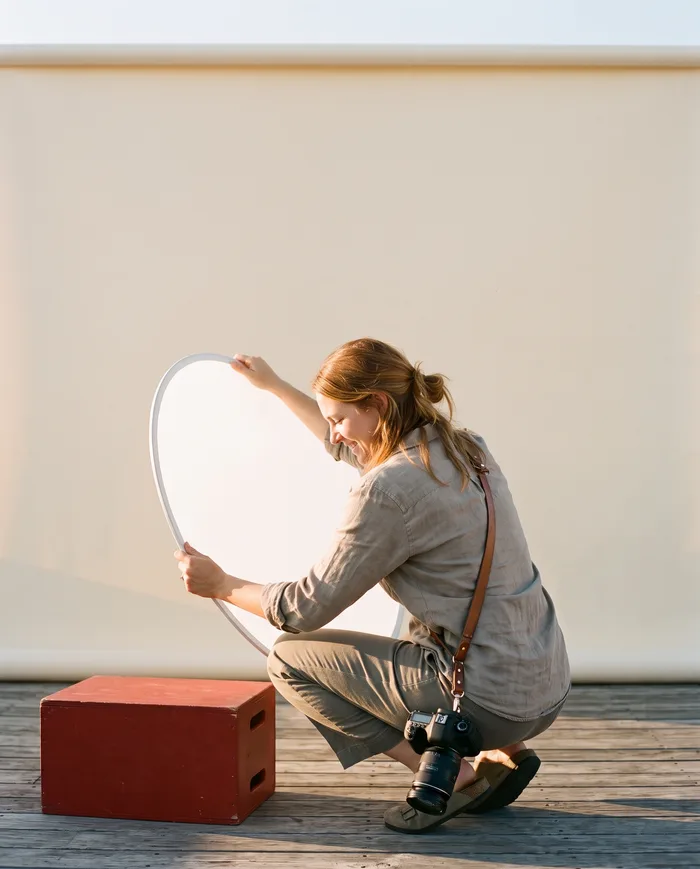 A woman crouched on a rooftop set in low golden-hour sun, angling a plain white reflector with both hands, a camera at her hip and a red crate on the deck